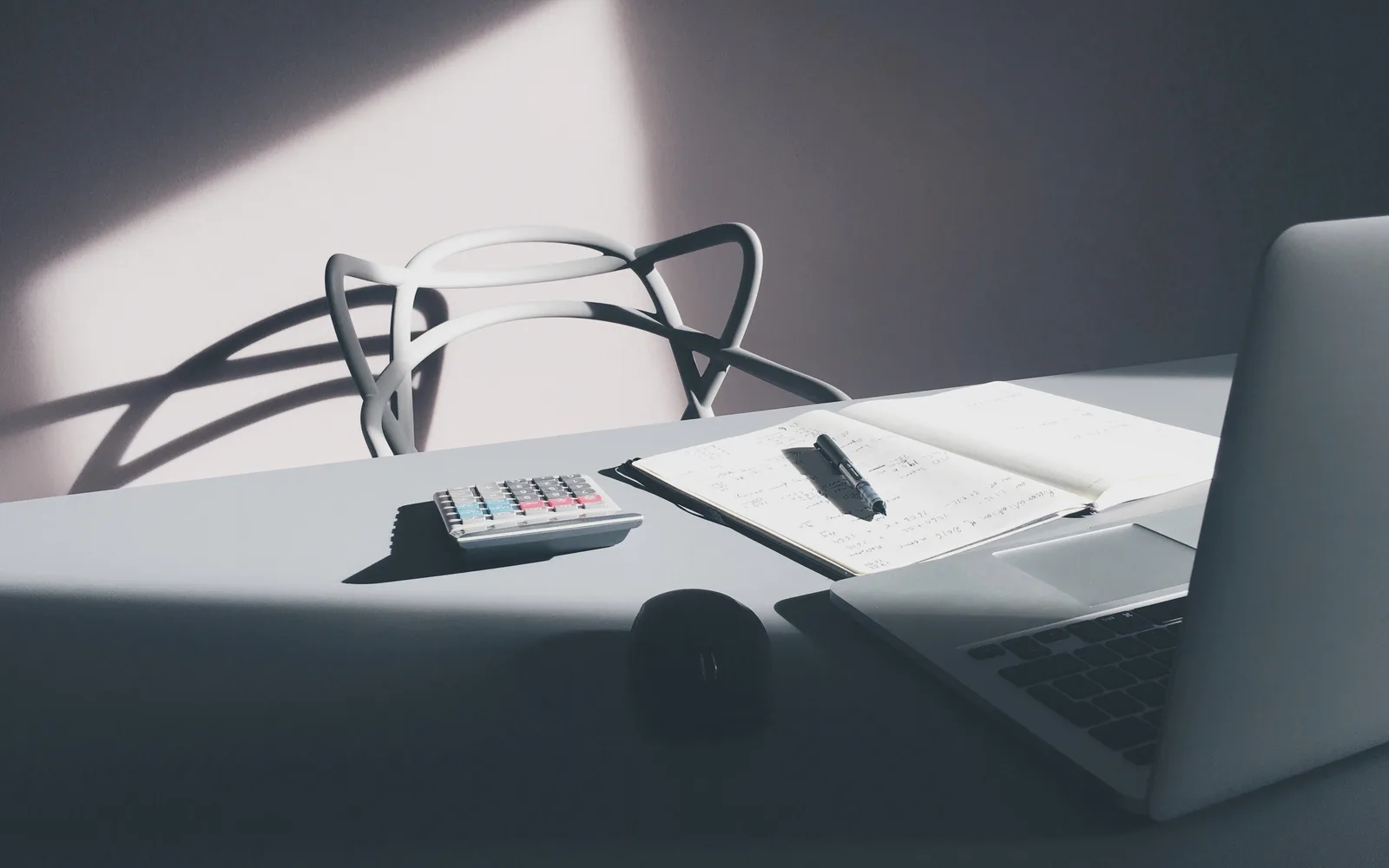 Calculator beside an open notebook with a pen and a laptop on a sunlit desk